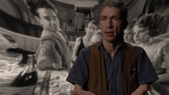 Image of Mat Fraser from Code of the Freaks. A white man wearing a denim shirt and a brown vest looks at the camera. The background image is a black and white still from Freaks (1932) with several people looking at the camera.