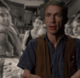 Image of Mat Fraser from Code of the Freaks. A white man wearing a denim shirt and a brown vest looks at the camera. The background image is a black and white still from Freaks (1932) with several people looking at the camera.