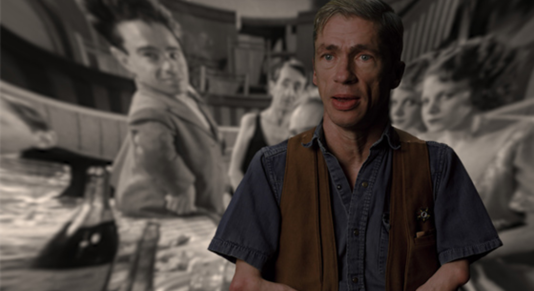 Still of Mat Fraser from Code of the Freaks. A white man wearing a denim shirt and a brown vest looks at the camera. The background image is a black and white still from Freaks (1932) with several people looking at the camera.