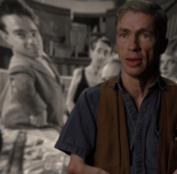 Still of Mat Fraser from Code of the Freaks. A white man wearing a denim shirt and a brown vest looks at the camera. The background image is a black and white still from Freaks (1932) with several people looking at the camera.
                  