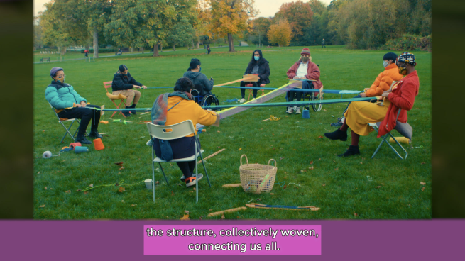 Eight people sit in a circle on a grassy field with green bushy trees in the background with soft lighting in the sky. Some sit in folded chairs and others in wheelchairs. Each person has a different gender expression, some people are trans and others are nonbinary. Most are people of color and there is one white person. All of the people pictured has a different disability. Some are wearing brightly colored jackets of light blues and bright oranges. Each person has warp threads tied around their bodies as backstrap looms connecting in crisscross shapes with lime green, purple, blue and orange threads. There are some weaving materials and yarn cones on the ground. At the bottom of the screen there are captions in white font on a purple pink background that read 