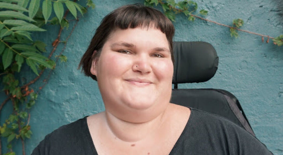 Kennedy, a white, fat, disabled femme, smiles with their head turned slightly to the right. They have short, brown hair and are wearing a black t-shirt. The back of their power chair is visible over their shoulder, and they are in front of a turquoise wall with green vines growing in the corner.