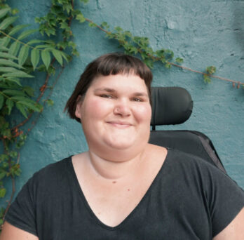 Kennedy, a white, fat, disabled femme, smiles with their head turned slightly to the right. They have short, brown hair and are wearing a black t-shirt. The back of their power chair is visible over their shoulder, and they are in front of a turquoise wall with green vines growing in the corner.
                  