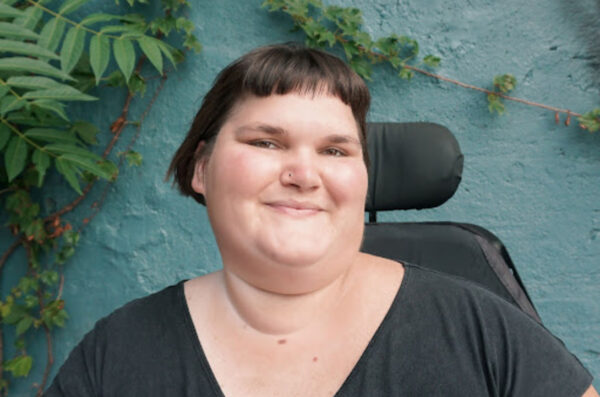 Kennedy, a white, fat, disabled femme, smiles with their head turned slightly to the right. They have short, brown hair and are wearing a black t-shirt. The back of their power chair is visible over their shoulder, and they are in front of a turquoise wall with green vines growing in the corner.