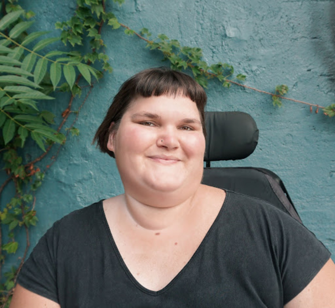 Kennedy, a white, fat, disabled femme, smiles with their head turned slightly to the right. They have short, brown hair and are wearing a black t-shirt. The back of their power chair is visible over their shoulder, and they are in front of a turquoise wall with green vines growing in the corner.