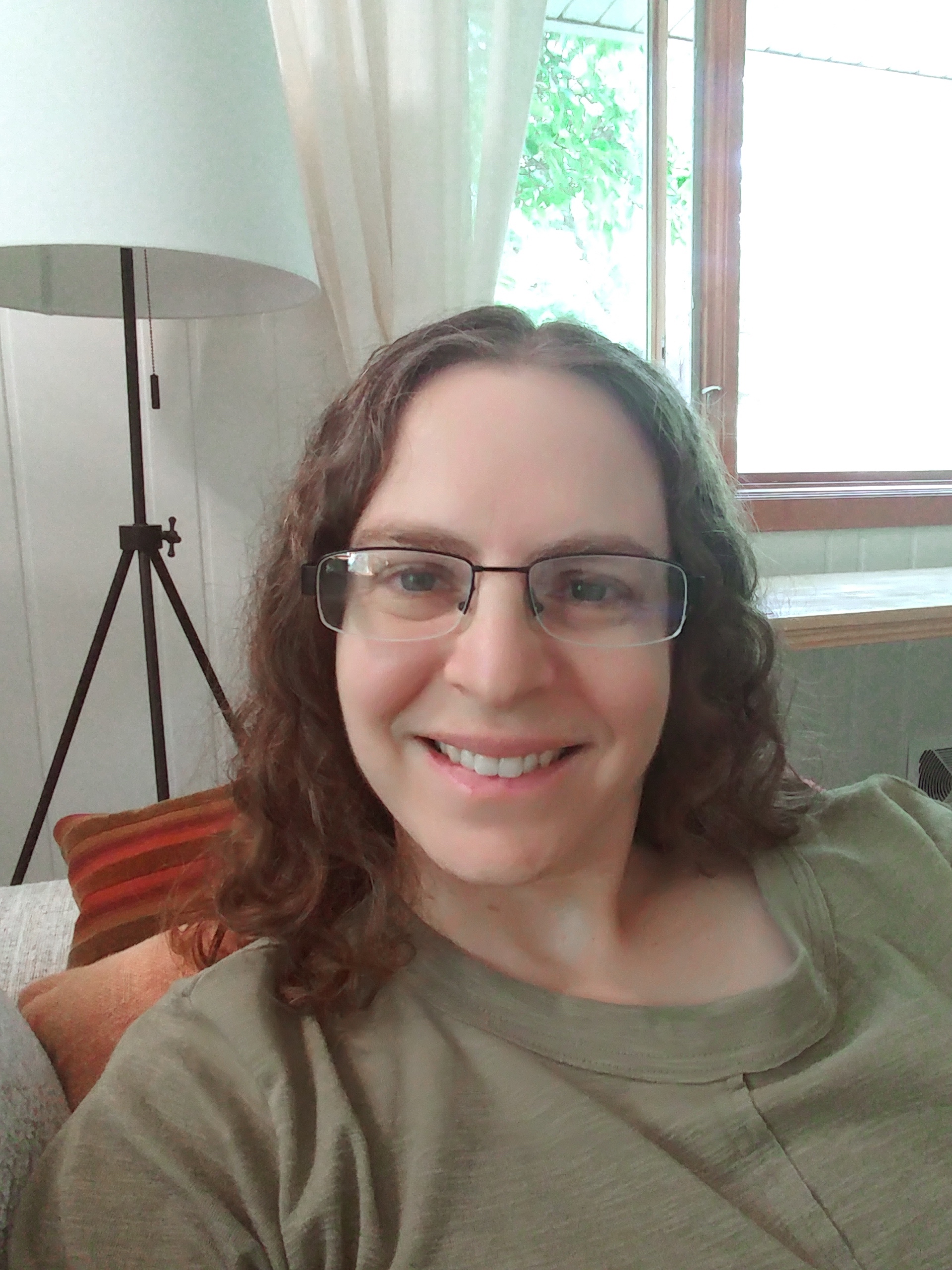 Alana is seated on a couch covered in pillows in front of an open window with light coming through. She wears rectangular glasses, an olive green t-shirt and her brown curly hair reaches her shoulders.