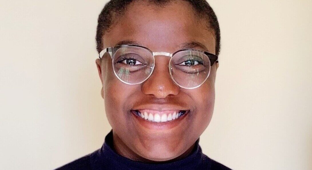 Nic, a Black person with round half-rimmed glasses, smiles at the camera. Nic wears a black turtleneck and is facing directly forward, framed against a light colored wall.