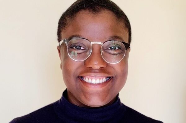 Nic, a Black person with round half-rimmed glasses, smiles at the camera. Nic wears a black turtleneck and is facing directly forward, framed against a light colored wall.
