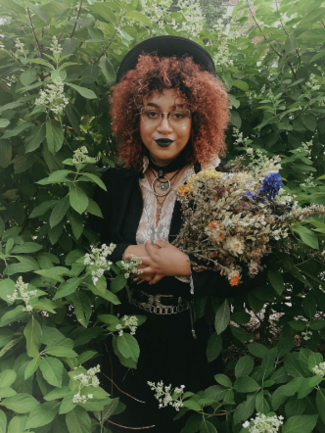 Bianca Xunise emerges from white floral bushes holding a bouquet. They are dressed in all black dress along with glasses and a hat.