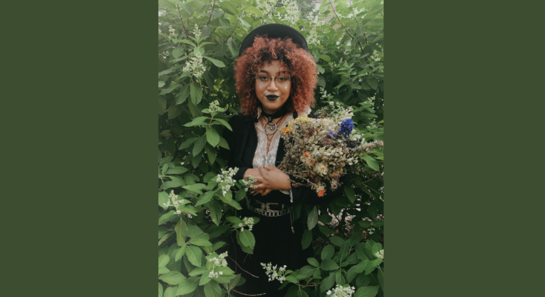 Bianca Xunise emerges from white floral bushes holding a bouquet. They are dressed in all black dress along with glasses and a hat.