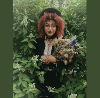 Bianca Xunise emerges from white floral bushes holding a bouquet. They are dressed in all black dress along with glasses and a hat.
                  