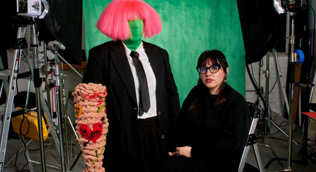 Ariella Granados, a femme Latinx artist in all black with glasses, long dark hair with bangs, and red lipstick, sits on a stool next to a green-screen fabric-clad mannequin wearing a pink wig with bangs and a suit and tie and a textured woven hand with patches made to look like tattoos including one that says “mom” in a heart with an arrow. In the background are a green screen,  lights, and camera equipment.