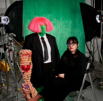 Ariella Granados, a femme Latinx artist in all black with glasses, long dark hair with bangs, and red lipstick, sits on a stool next to a green-screen fabric-clad mannequin wearing a pink wig with bangs and a suit and tie and a textured woven hand with patches made to look like tattoos including one that says “mom” in a heart with an arrow. In the background are a green screen,  lights, and camera equipment.
                  