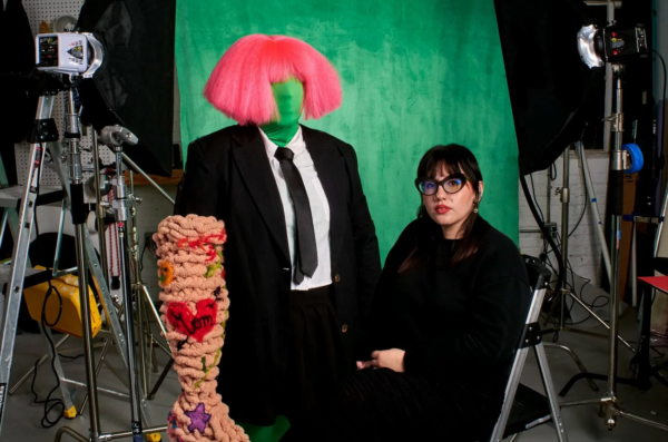 Ariella sits in a chair dressed in black on the right side of the image with a green screen and lighting equipment behind them. They have their dark hair pulled back with straight bangs and are wearing bright red lipstick. To the left of Ariella is a mannequin with a green face, bright pink hair and one giant crocheted arm with tattoos.