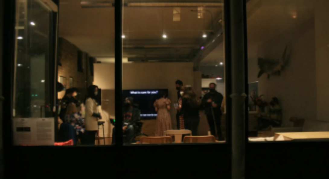 A picture of Curb Appeal’s inaugural opening of Molly Joyce’s exhibit Prescriptive. This photo is taken from the outside looking through the window of the apartment art gallery. Inside, people are standing in front of chairs. Behind the people is a large television screen displaying Joyce exhibit Prescriptive. The screen is black and the top of the screen reads “What is cure for you?” in white letters. Photo courtesy of Curb Appeal Gallery.