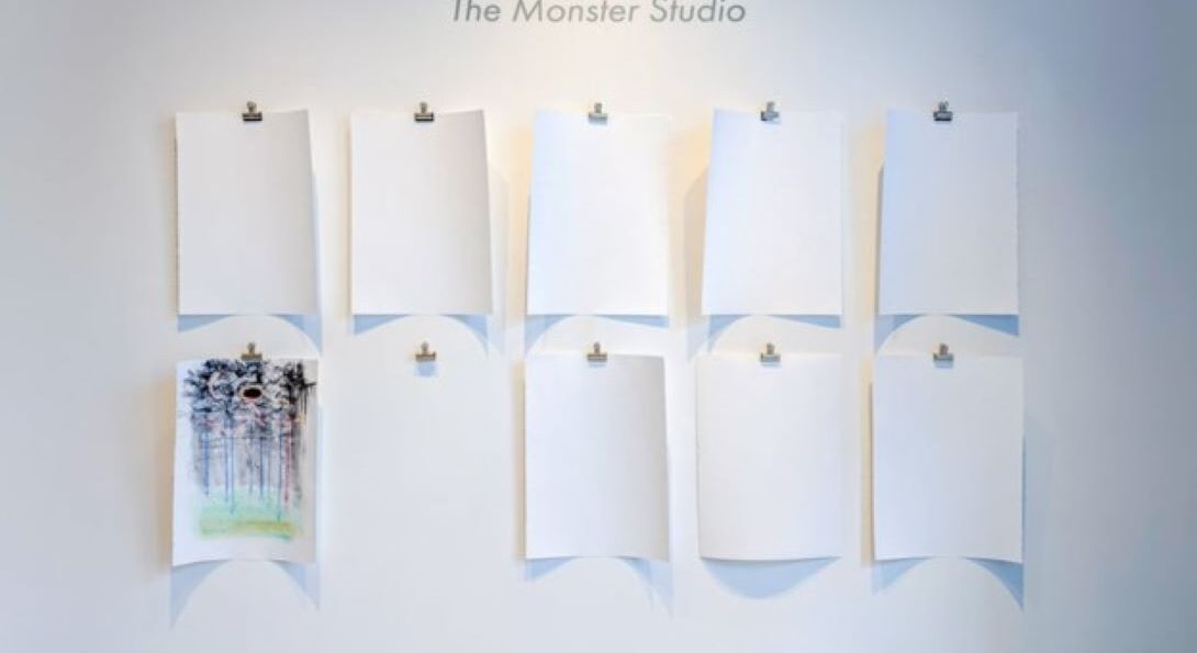 A white wall contains the text: RIVA LEHRER The Monster Studio. The wall has nine pieces of white paper in two rows hanging from silver bulldog clips. All the paper is blank except for one that contains a chromatic, abstract drawing. There is an empty bulldog clip where the next completed drawing will hang.